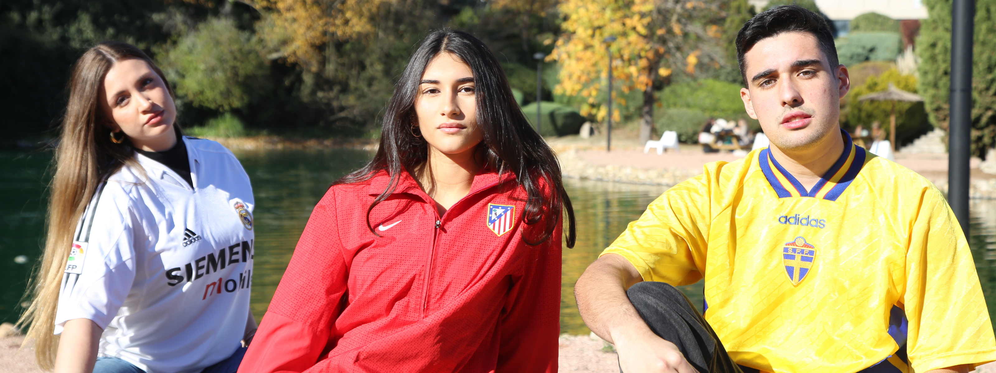 Dos mujeres, una con la camiseta del Real Madrid y otra con una sudadera del Atlético de Madrid, sentados al borde de un lago con un chico con una camiseta retro de la Selección de Futbol de Suecia. 